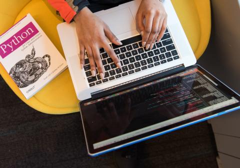 Photo by Christina Morillo. Girl at computer with a book about the code Python next to her. 